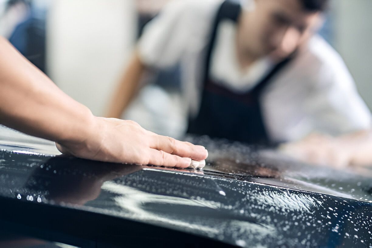 Focus on hand of worker holding film. Detailing of two Asian car film protection male worker installing film to black color car in garage shop.